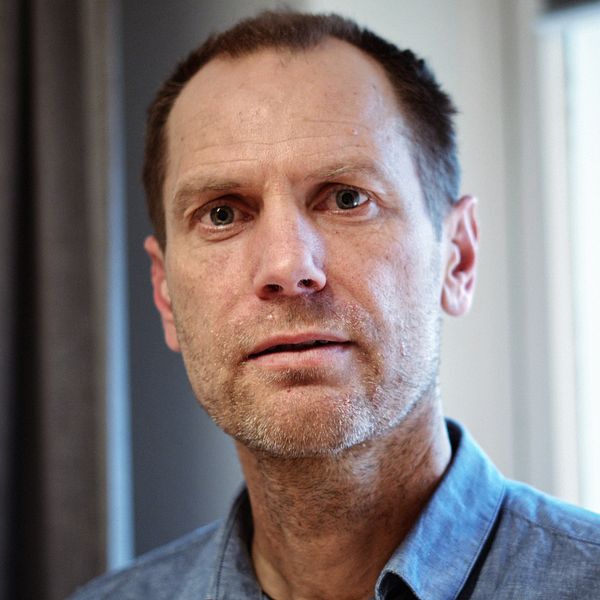 En person står inomhus nära ett fönster iklädd en jeansskjorta, med gardiner synliga i bakgrunden.