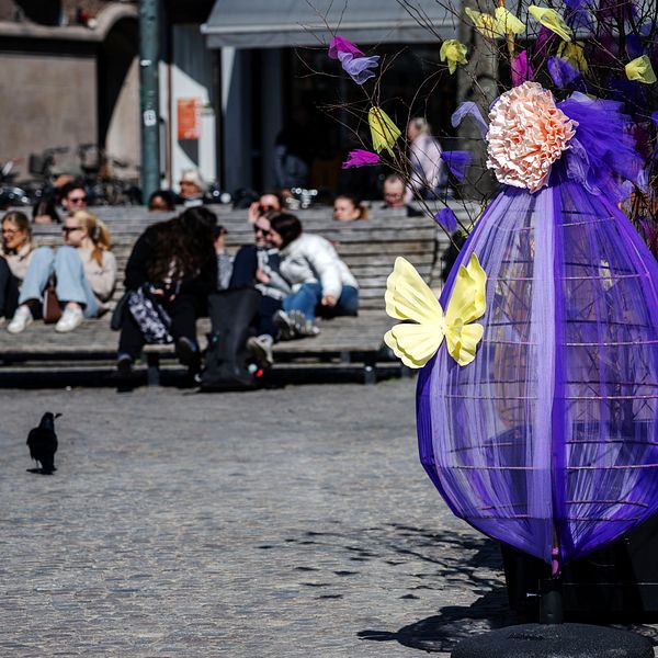 Utsmyckat påskträd på torg. Människor sitter på bänkar i bakgrunden.