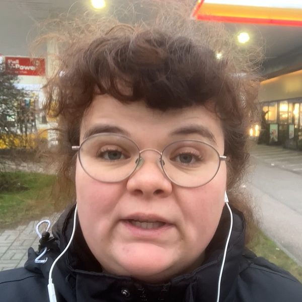 Reporter Klara Misgeld står utomhus i en stadsmiljö nära en bensinstation, med en byggnad i bakgrunden som tyder på kommersiell verksamhet. Det är en grå och mulen dag. Personen har hörlurar och bär en svart jacka. Det finns parkering i bakgrunden tillsammans med buskar och en del av en gångväg.
