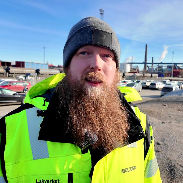 Tomas Hart med lysande grön jacka står vid industriområde med bilar och fabriker i bakgrunden.