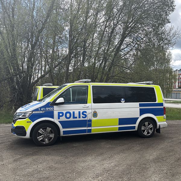 En svensk polisbil står parkerad vid en väg nära träd och en bro över vatten.