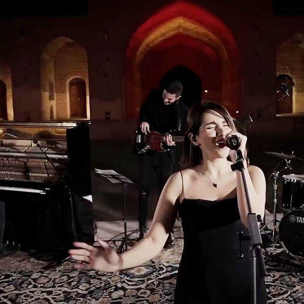 Parastoo Ahmadi i Iran framträder på en scen utomhus, omgiven av en pianist, en gitarrist och en trummis. Bakgrunden har vackert upplysta bågar och väggar.
