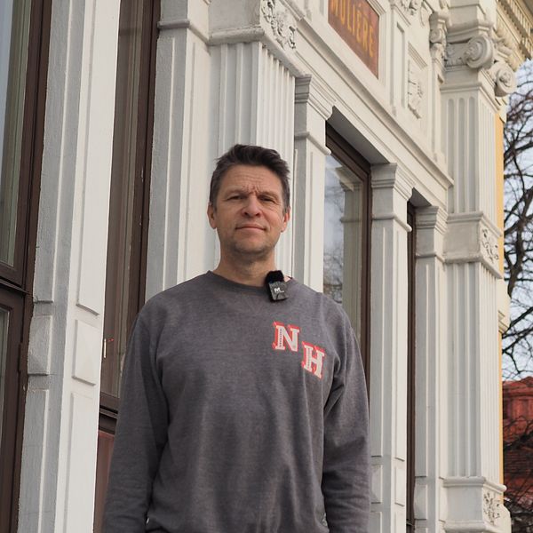 En person står utanför en byggnad med klassisk arkitektur, inklusive dekorerade fönster och detaljerad fasad. Bakgrunden visar träd utan löv, vilket ger en vinterliknande atmosfär. Personen bär en gråtröja med ett rött mönster på bröstet.