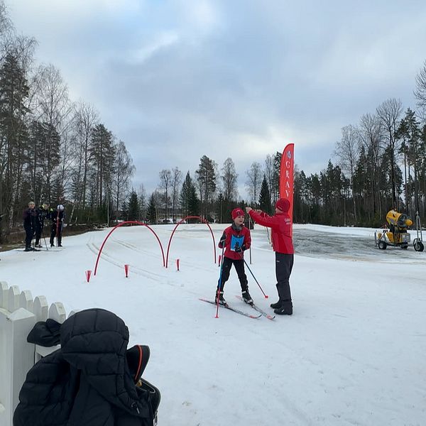 En skidtränare i röd jacka ger instruktioner till en ung skidåkare i blå och röd utrustning vid en skidbana. Bakgrunden visar människor som tittar på och en snöskoter som preparerar spåret. Tränaren står vid en mållinje markerad med röda portaler.