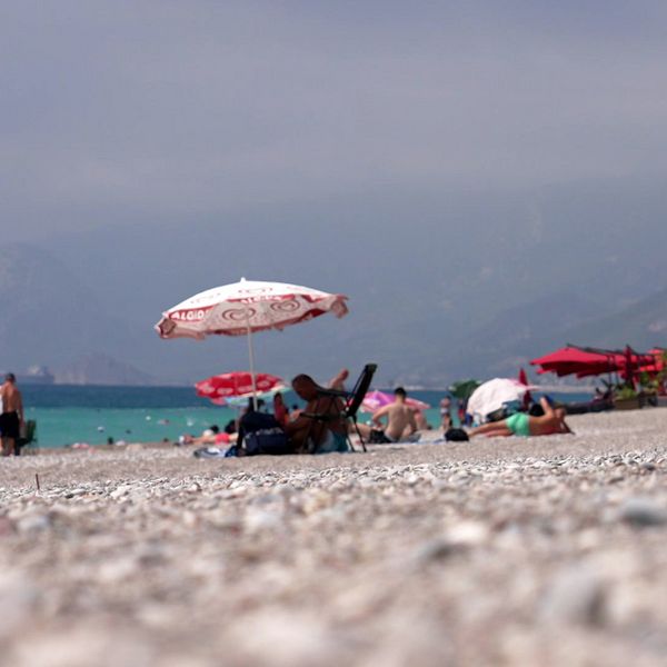 Turister vid populär strand med parasoller och berg i bakgrunden.