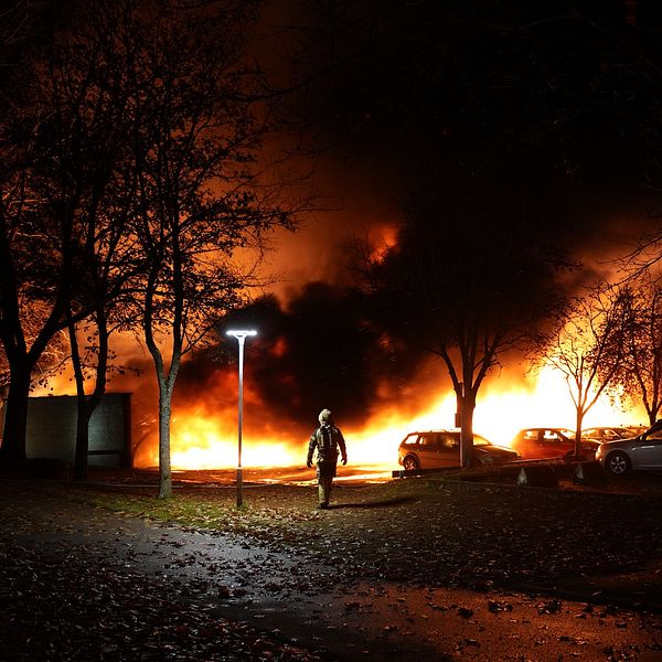 En person går genom en parkering i skymningen, med en stor brand som rasar i bakgrunden. Flammor och rök lyser upp natten, medan träden och bilarna ger en kontrast till den dramatiska scenen.