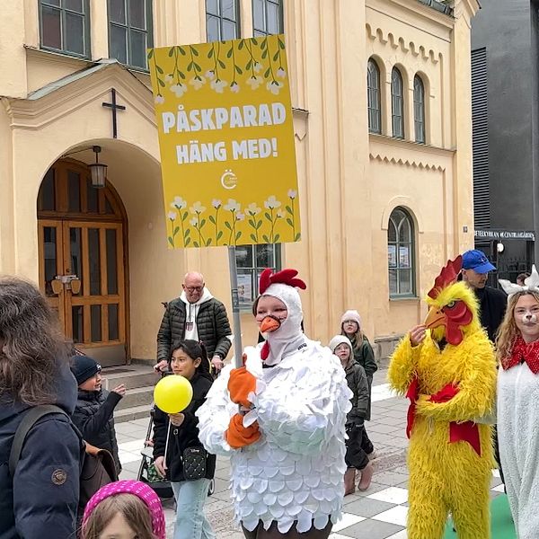 Bilden fångar en livlig scen på en stadsgata där människor, inklusive barn, samlas för ett evenemang. Tre personer är klädda i färgglada dräkter som liknar höns eller fåglar, och stämningen är festlig. En stor banderoll med texten ”PÅSKPARAD HÄNG MED!” visas på en byggnad och inbjuder människor att delta i påskparaden.