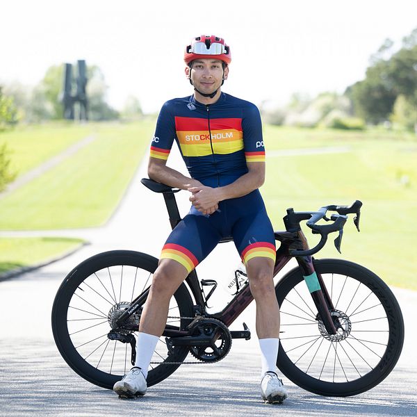 En cyklist i en färgglad cykeltröja och shorts sitter på sin racingcykel i en park. Det är en solig dag med grönskande omgivningar och en välskött väg i bakgrunden. Cykeln har en modern design med slanka hjul och aerodynamiska detaljer.