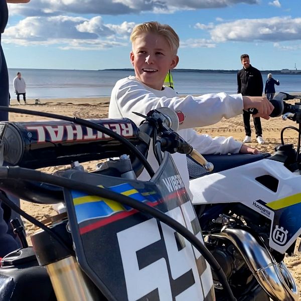 En pojke sitter på en motorcrosscykel på en sandstrand, omgiven av flera personer som står i bakgrunden. Det är en solig dag med några moln på himlen och havet syns i fjärran. Cykeln i fokus är vit med blå och gula detaljer, medan en annan motorcykel med nummer 54 också syns i bildens främre del.