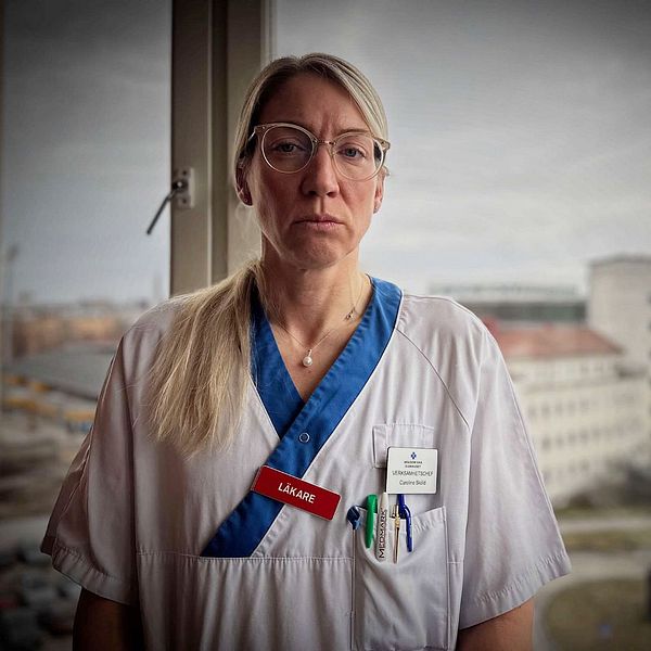 Sjukvårdspersonal i vit uniform står vid fönster med sjukhusbyggnader i bakgrunden.