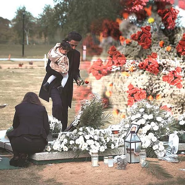 En familj samlas vid en gravplats för att hedra en avliden person. Två personer står vid graven, där en man håller ett barn, medan en kvinna sitter på knä vid gravstenen. Omkring graven finns det en stor uppsättning blommor, mestadels vita, tillsammans med ljus och andra minnesföremål. I bakgrunden syns en närbild av coronaviruset.