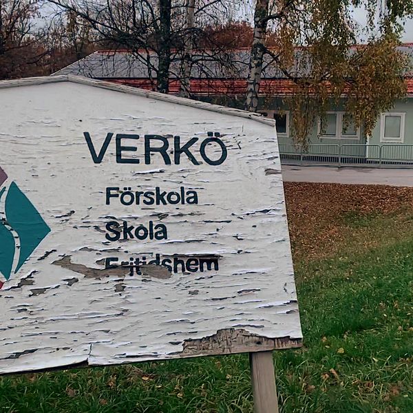 Skylt vid Verkø Förskola, Skola och Fritidhem med en suddig bakgrund av byggnaden och omgivande träd i höst. Skyltens text visar namnet på institutionen och dess verksamheter.