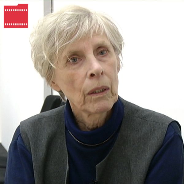 En person med grått hår sitter på en stol i ett ljust rum, klädd i en mörkblå tröja och en väst. Rummet har en minimalistisk inredning med vita väggar och svarta stolar i bakgrunden.