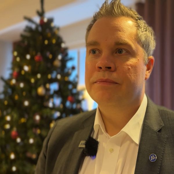En person står framför en julgran dekorerad med färgglada kulor och ljus. Bakgrunden visar ett rum med gardiner i dämpade färger och ett fönster som släpper in naturligt ljus. Personen bär en mörk kostym med en vit skjorta och har en mikrofon fäst vid kragen.