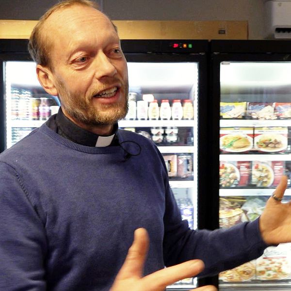 En person i en blå tröja och prästkrage gestikulerar framför ett kylskåp fyllt med matvaror och drycker i en affär eller butik.