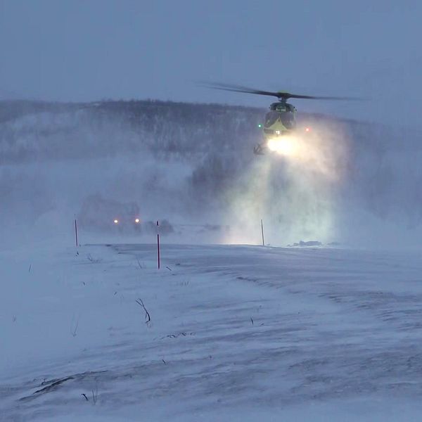 Helikopter lyfter i vinterlandskap, virvlande snö och dimma i bakgrunden.