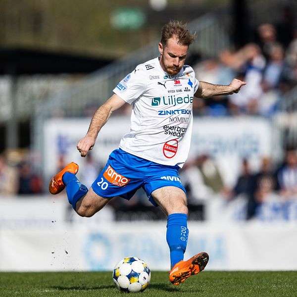 Fotbollsspelare sparkar boll under match på gräsplan med publik i bakgrunden.