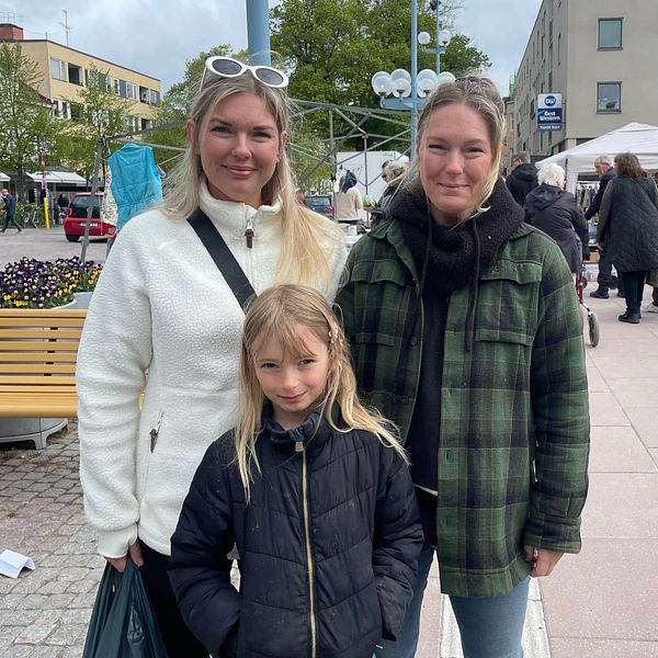 Tre personer står på ett torg, marknadsstånd och personer i västar syns i bakgrunden.