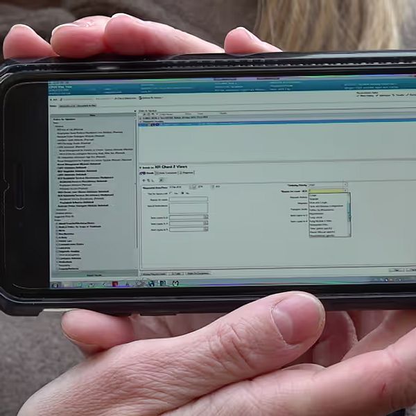 En person håller upp en smartphone som visar en skärm med programvara, med olika alternativ och fält för information. Skärmen verkar vara en del av ett gränssnitt för datainmatning eller hantering.