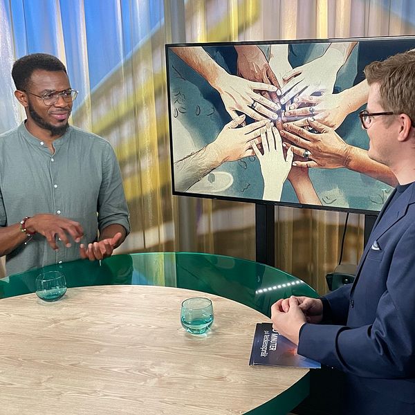 Romel Belcher och Magnus Bergevin står vid ett runt bord i en studio. I bakgrunden visas en tv-skärm en illustrerad bild med flera händer som symboliserar samarbete och gemenskap. På bordet finns två vattenglas.