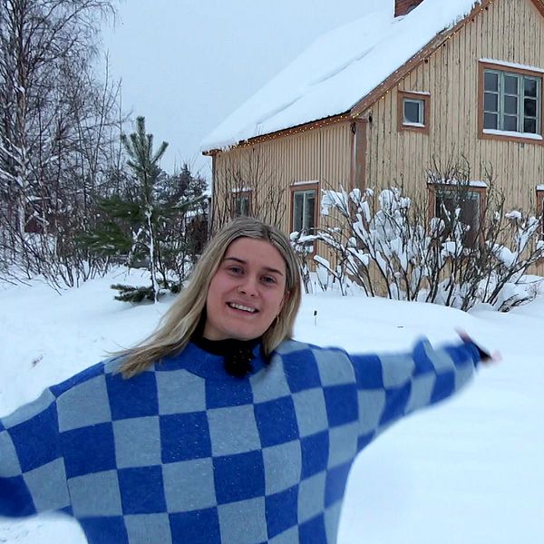 Anna Nilsson har en blårutig tröja och står med armarna utsträckta framför huset hon köpt i Gargnäs. Det syns att huset stått tomt en längre tid, med stora färgflagor i den beiga färger och de bruna husknutarna. Det är vinter och snö på marken.