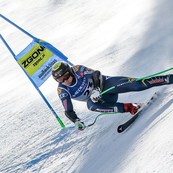 En alpin skidåkare i full fart svänger genom en blå port under en slalomtävling på en snötäckt backe. Skidåkaren bär en mörk dräkt med olika sponsormärken och är fokuserad på att manövrera genom banan.