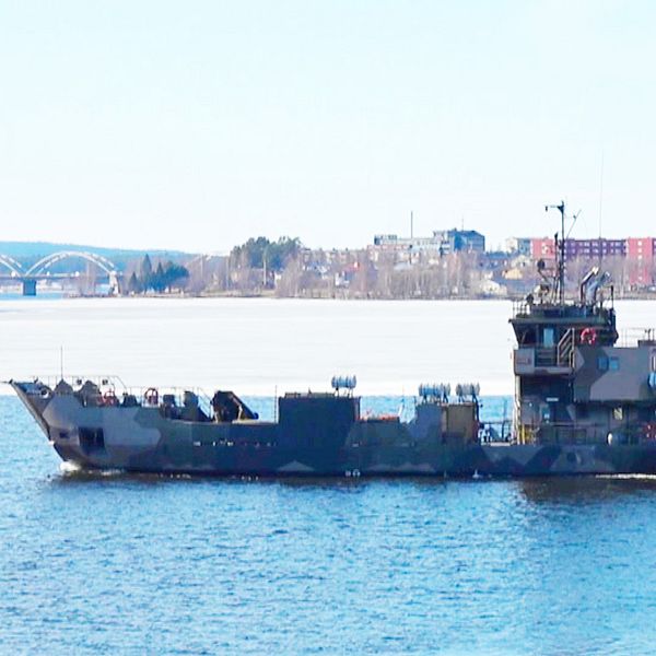 Militärfartyg färdas på vatten med stad och bro i bakgrunden.