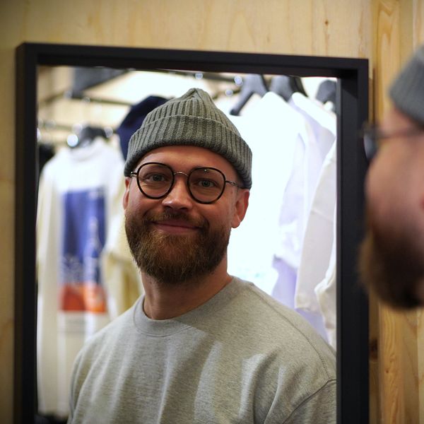 En person i en grå tröja och en grå beanie står framför en spegel i en klädaffär. Bakgrunden visar klädesplagg som hänger på galgar, vilket skapar en butiksmiljö.
