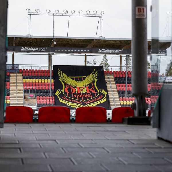 Stor svart banner med Östersunds klubbemblem hänger framför tomma läktare på en fotbollsarena, med rader av röda och gula stolar synliga. Bilden fångar en vy från en ingång till arenan, vilket ger en känsla av förväntan inför en match.