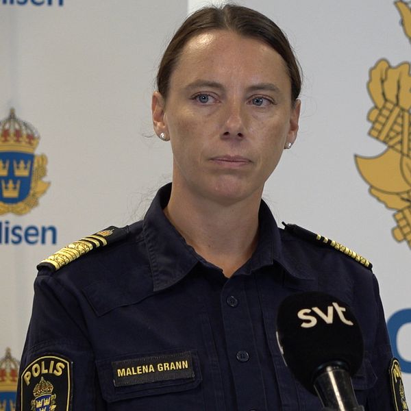 Kvinnlig polis står i uniform framför vit vägg med polisens logotyper på.