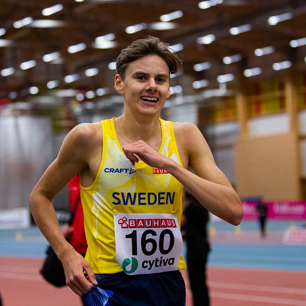 En löpare i tävlingskläder med texten ”SWEDEN” och nummerlappen 160 springer inomhus på en löparbana. I bakgrunden syns tränare och andra deltagare i en stor hall med träinredning och neonljus.