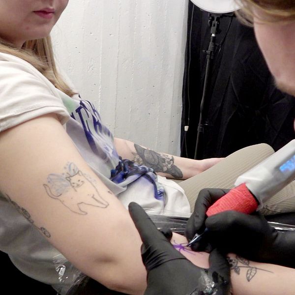 En person sitter i en tatueringsstudio medan en tatuerare arbetar med att fullfölja en tatuering på hennes arm. Armens tatueringar är synliga, inklusive en design som föreställer en katt. Tatueraren använder en maskin med röd färg. Omgivningen är professionell och inbjudande.