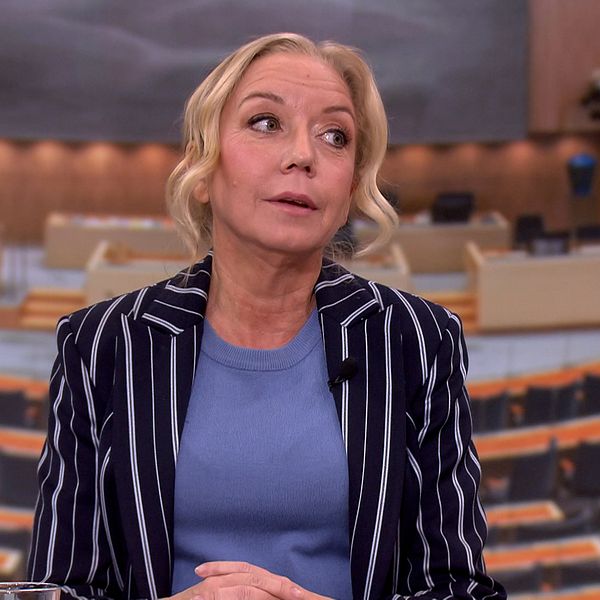 Kvinna i kavaj framför en bakgrund av riksdagens plenisal.