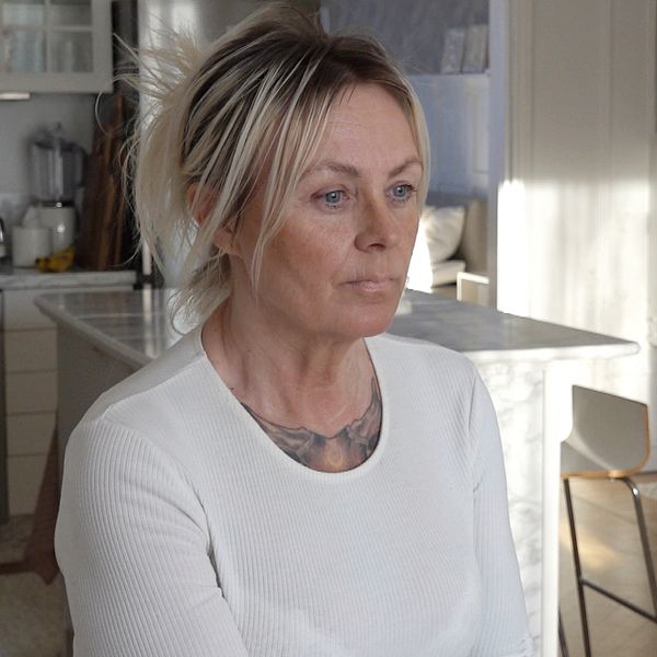Kvinna i vit långärmad tröja sitter i ett modernt kök med vita marmordetaljer och trädetaljer i bakgrunden.