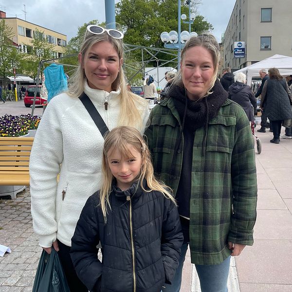 Tre personer står på ett torg, marknadsstånd och personer i västar syns i bakgrunden.