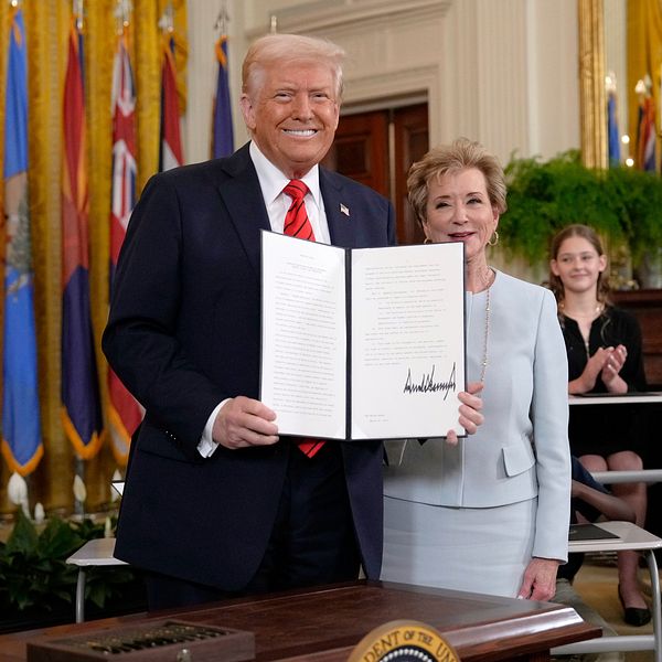 Dobald Trump håller upp ett dokument. I bakgrunden ser man flaggor och applåderande personer.