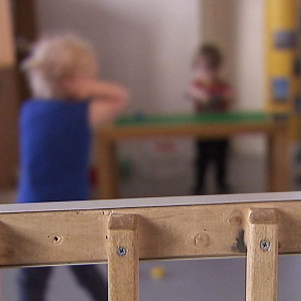 Interiör av en förskola med små barn som leker i bakgrunden och en träbarriär i förgrunden. Barnen är upptagna med olika aktiviteter i ett rymligt rum.