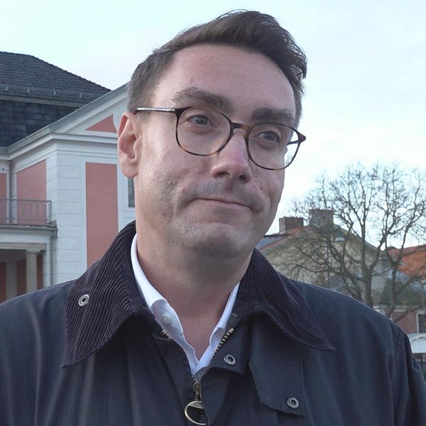 En person står framför en historisk byggnad med vit och rosa fasad. I bakgrunden syns träd och andra hus, vilket skapar en stadsbild. Personen bär en mörk jacka och en vit skjorta. Himmelns nyanser är ljusa med några moln.