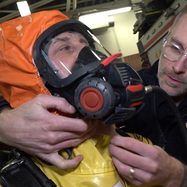 Brandmän hjälper varandra med att sätta på sig utrustning för att kemdyka. En man hjälper en anna med att sätta på dig en orange galonhätta som ska skydda mot giftiga kemikalier. Mannen med hätta har en stor ansiktsktsmask på sig.
