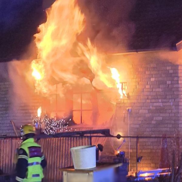 Ett tegelhus täckt av lågor under en brand, med en brandman i gul skyddsutrustning i förgrunden som verkar vara på väg mot eldsvådan. Elden ryker intensivt från husets fönster.