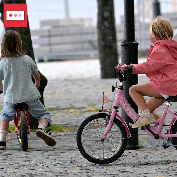 Två barn cyklar på en grusväg; ett i rosa jacka och ett med långt hår. Ett barn står bredvid.