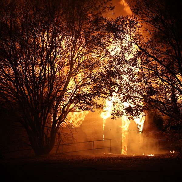Ett hus omgärdat av stora lågor som sprider sig kraftigt, med träd som skymmer en del av byggnaden. Scenen präglas av en intensiv brand som lyser upp omgivningen i orangeröda och gula nyanser. Rök kan också ses i luften runt omkring.