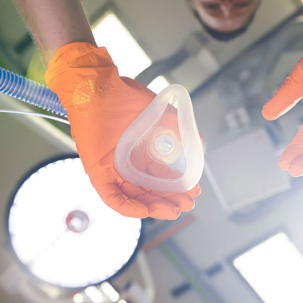 En person med orangea handskar håller en anatomisk respiratormask i handen, medan de ser ner mot den. I bakgrunden syns svagt upplysta kirurgiska lampor och en del av en medicinsk miljö, vilket tyder på att bilden är tagen i en operationssal eller liknande.