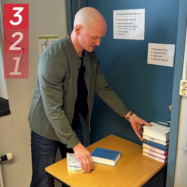 Säkerhetssamordnaren Johan Leopoldsson, i en grön jacka, står i ett rum och placerar böcker på ett litet bord under handtaget på en blå dörr.