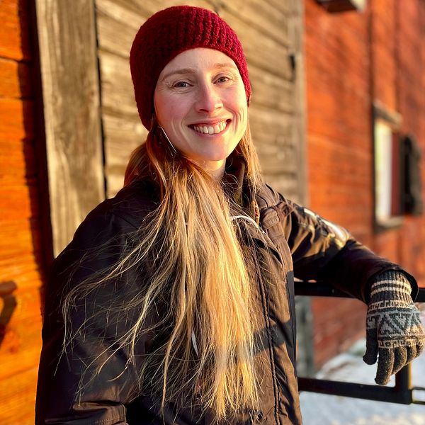 En person står lutad mot ett räcke framför en gammal träbyggnad med rödbrun fasad. Personen har på sig en mörk jacka och en röd mössa, och deras lång hår hänger ner över axlarna. I bakgrunden syns snö och träd, vilket tyder på kallt väder. Solens strålar belyser scenen och skapar en varm atmosfär.