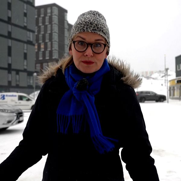Person står utomhus på en snötäckt gata, med moderna byggnader i bakgrunden.