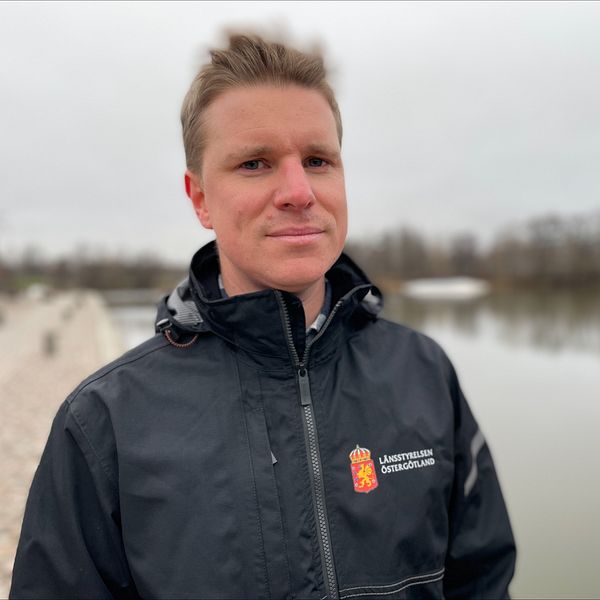 En person står vid vattnet med en mörk jacka som har en logotyp på bröstet. I bakgrunden syns en stilla sjö och träd längs strandlinjen under en grå himmel.