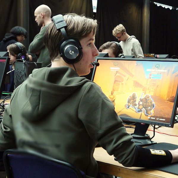 En grupp personer i en spelturnering, där fokus ligger på en spelare som sitter vid en dator med hörlurar. Skärmen visar en actionsekvens från ett videospel. Omkring finns andra deltagare och en publik som observerar evenemanget. Atmosfären är livlig och fokuserad på spelande.