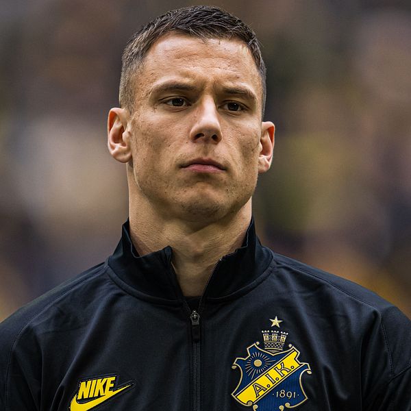 AIK-spelare med mörkt jacka och klubbmärke synligt, suddig publik i bakgrunden.
