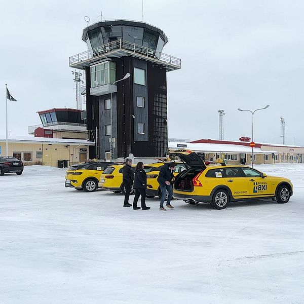 En vinterbild av Örnsköldsviks flygplats med snö på marken och flygledartornet i bakgrunden. Flera gula taxibilar står parkerade i förgrunden där några personer lastar bagage ur en taxi.